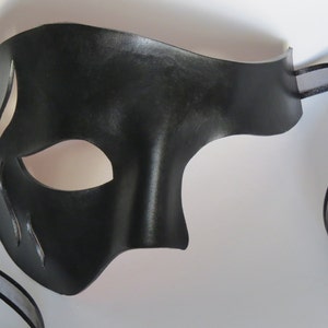 May include: A black leather masquerade mask with a single eye opening and a decorative cut-out design. The mask is attached to a black ribbon.