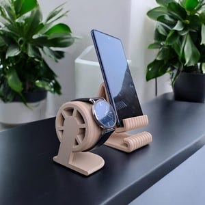 Könnte beinhalten: Ein hellbeige Holzständer für Telefon und Uhr. Er hält ein schwarzes Smartphone und eine Armbanduhr mit schwarzem Lederarmband und blauem Zifferblatt. Der Ständer steht auf einer dunklen Oberfläche.