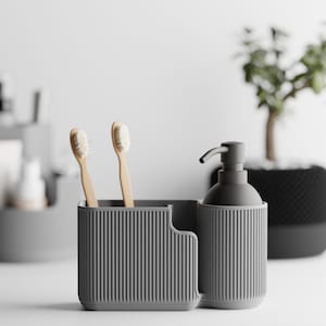 Könnte beinhalten: Graues Badezimmer-Organizer-Set mit Zahnbürstenhalter, Seifenspender und kleiner Pflanze im Hintergrund. Der Zahnbürstenhalter hat zwei Zahnbürsten. Das Set besteht aus geripptem Material.