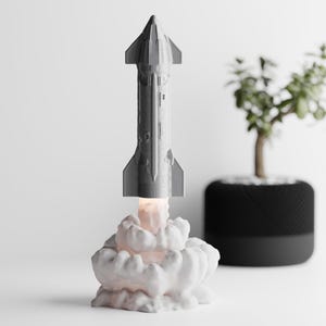 Peut inclure: Lampe en forme de fusée grise avec une base en forme de nuage blanc, émettant une lueur chaude. La lampe est une maquette détaillée d'une fusée, avec une petite plante en arrière-plan.