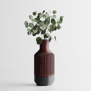 May include: A maroon ceramic vase with vertical ribbed detailing and a dark gray base. The vase holds a collection of green eucalyptus leaves. The vase is a decorative item for home decor.