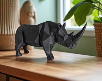 May include: A black geometric rhino sculpture with a long horn stands on a wooden surface. The rhino is made of many flat, angled surfaces, giving it a modern, angular appearance. The background includes plants and a window.