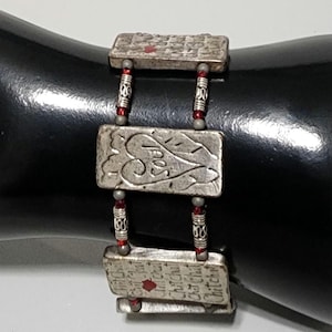 May include: A multi-strand bracelet featuring three rectangular silver-toned metal plates with engraved text and heart motifs. The plates are connected by small silver-toned cylindrical beads and tiny red glass beads, creating a layered design.