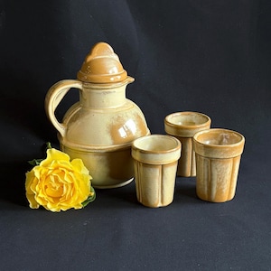 May include: A ceramic pitcher with a lid and three matching tumblers, all featuring a mottled tan and cream glaze. The pitcher has a curved handle and a spout. A single yellow rose is positioned in front of the pitcher on the dark surface.
