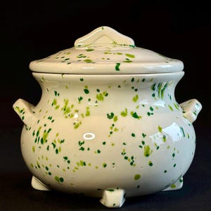 May include: A white ceramic lidded pot with green speckled detailing. The pot has two small handles and three feet. The lid has a small handle. The pot is likely used for storage or decoration.