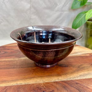 May include: A handmade ceramic bowl with a dark brown exterior and a glossy black interior. The rim has a lighter brown color. The bowl sits on a wooden surface.