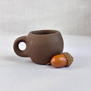 May include: A small, brown ceramic mug with a rounded body and a circular handle. Beside the mug is a realistic acorn with a brown cap and an orange-brown nut. The mug and acorn are set against a white background.