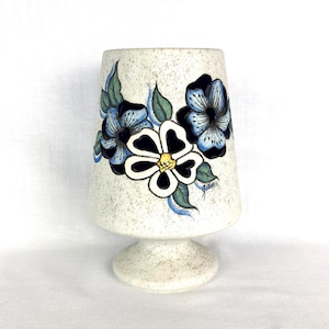 May include: A vintage, off-white ceramic vase with a speckled finish. The vase features hand-painted blue and white flowers with green leaves. The vase has a flared top and a pedestal base, showcasing a retro floral design.