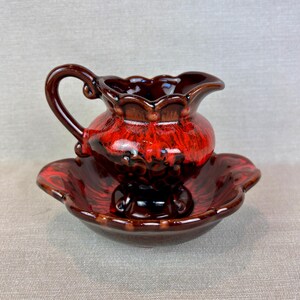 May include: A vintage ceramic pitcher and basin set. The pitcher is a deep red with a glossy finish and a decorative handle. The basin is a matching red with a scalloped edge. The set is likely for decorative use.
