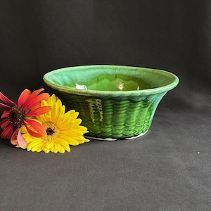 May include: A glossy green ceramic bowl with a textured basket-weave pattern on the exterior and a smooth interior. The bowl is positioned next to a vibrant red coneflower and a bright yellow daisy, creating a contrast of natural floral colors.