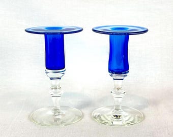 May include: Two cobalt blue glass candle holders. Each has a round base, a clear stem, and a vibrant blue top. The candle holders are displayed against a white background, highlighting their elegant design. These decorative pieces add a touch of color and sophistication.