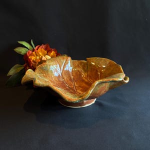 May include: A ceramic bowl shaped like a large leaf with a glossy, mottled finish in shades of amber, brown, and green. The bowl features organic, wavy edges and visible vein-like indentations on its surface, resting on a small circular base.