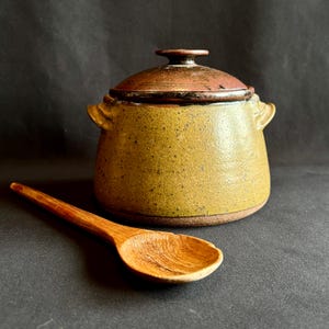 May include: A round ceramic pot with a textured mustard-yellow body, two small side handles, and a dark reddish-brown lid. The pot features a speckled finish and is accompanied by a long-handled wooden spoon with a visible natural grain pattern.