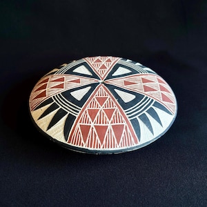May include: A shallow, circular ceramic bowl featuring a geometric pattern in terracotta, black, and white. The design consists of repeating triangles and radiating lines that converge at the center, creating a symmetrical, intricate visual motif.