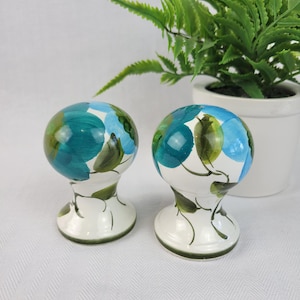 May include: A pair of ceramic salt and pepper shakers. Each shaker has a white base with a green rim and a spherical top. The tops are decorated with teal, blue, and green floral designs.