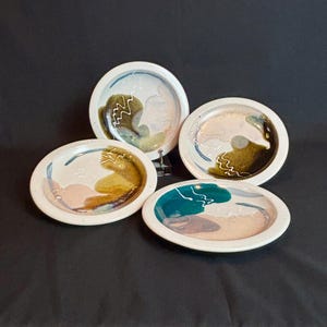 May include: Four ceramic plates with abstract designs. The plates have a white base with colorful accents of teal, brown, pink, and blue. Each plate features a unique pattern with a wavy white line design.