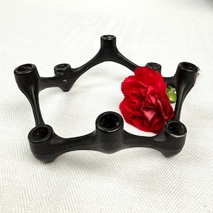 May include: A black metal candle holder with six circular openings for candles. The holder has a unique, curved design and is accompanied by a vibrant red flower. The piece is set against a white background.