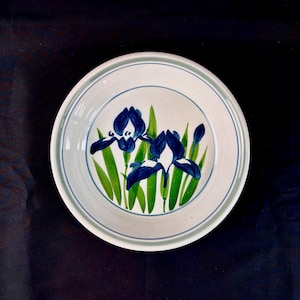 May include: A white ceramic plate with a blue and green floral design. The plate features two blue iris flowers with green leaves and a blue border. The plate is round and has a simple, elegant design.