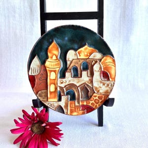 May include: A round ceramic wall plaque featuring a relief design of a Middle Eastern cityscape with domes and a minaret. The piece is glazed in shades of deep blue, cream, orange, and brown, with the artist signature SAR visible in the lower left corner.