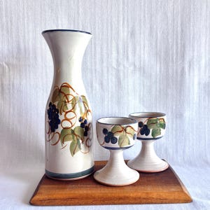 May include: A tall ceramic carafe and two matching stemmed goblets featuring a hand-painted design of dark blue grapes, green leaves, and brown swirling vines on a cream-colored background with dark blue rim accents on each piece.