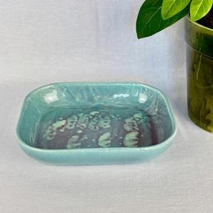 May include: A rectangular ceramic dish with a turquoise glaze. The interior features a mottled pattern of teal and purple. The dish has rounded corners and a slightly raised rim. A green plant in a green pot is in the background.