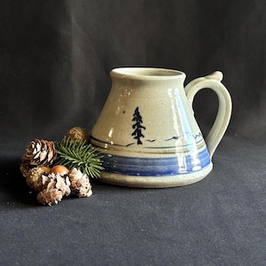 May include: A wide-based ceramic mug with a speckled off-white glaze, featuring a dark blue painted band at the base and a small black pine tree illustration. The mug has a curved handle and is accompanied by a cluster of small brown pinecones.