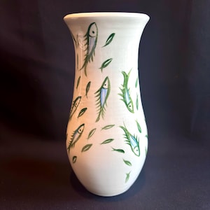 May include: A white ceramic vase with a flared top, hand-painted with green and blue fish and green leaf designs. The vase is approximately 10 inches tall, with a smooth, glossy finish. A decorative piece for home decor.