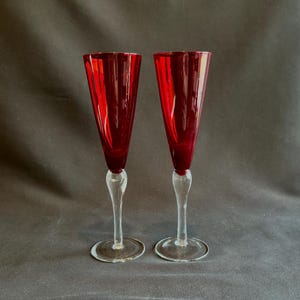 May include: Two elegant champagne flutes with ruby red bowls and clear stems and bases. The flutes have a classic, tapered shape and are set against a dark background. Perfect for a special occasion.
