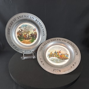 May include: Two pewter plates with colorful historical scenes from the American Revolution. The plates are round with the text "THE GREAT AMERICAN REVOLUTION" and the year "1776" around the edges. One plate depicts a battle scene, the other a boat crossing.