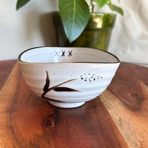 May include: A white ceramic bowl with a brown rim and a decorative black floral design. The bowl has a square shape with rounded corners and is sitting on a wooden surface. The bowl has three "X" marks on the rim.