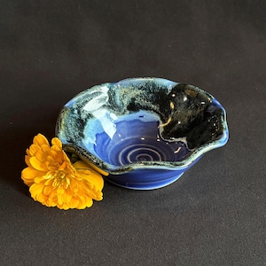 May include: A small, round ceramic bowl with a wavy, scalloped rim. The interior features a deep blue base color that transitions into a dark, mottled black and teal glaze along the edges. A bright yellow flower is positioned next to the bowl.