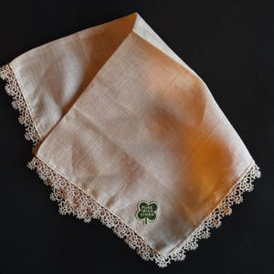 May include: A folded, peach-colored linen handkerchief with a delicate lace trim. The handkerchief features a green emblem with the words "Pure Irish Linen" and a shamrock design. The fabric has a subtle sheen, and the lace adds a touch of elegance.