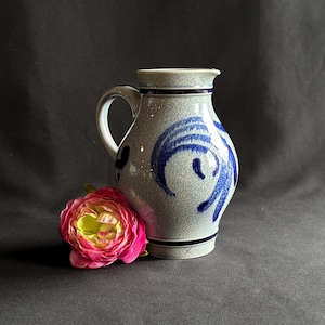 May include: A ceramic pitcher with a speckled light gray glaze and hand-painted dark blue swirling patterns. The vessel features a curved handle, a flared rim, and two horizontal dark blue bands. A pink and yellow artificial flower sits nearby.