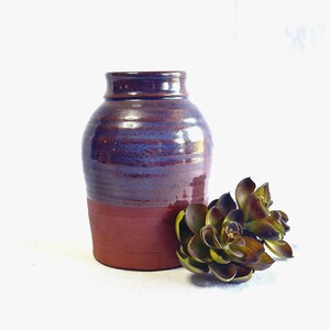 May include: A handcrafted ceramic vase with a two-tone design. The upper portion is a deep purple with blue hues, while the lower section is a rich, earthy red. A decorative succulent plant sits beside the vase.