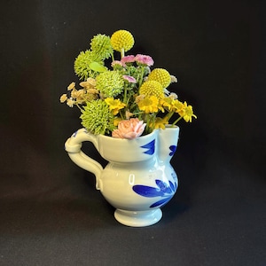 May include: A white ceramic pitcher with a handle and blue floral designs, filled with a variety of colorful flowers. The flowers include yellow, green, pink, and peach blooms. The pitcher is on a black background.