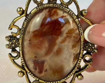 May include: Vintage oval brooch with a gold-tone filigree frame. The brooch features a large, polished stone with a mix of brown, cream, and reddish-brown hues. The frame has ornate details and small, round accents.