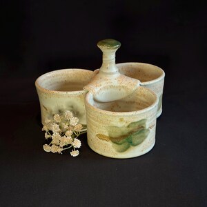 May include: A handmade ceramic condiment set with a lid and two open compartments. The set is beige with green and brown accents. A small bouquet of white flowers sits next to the set.