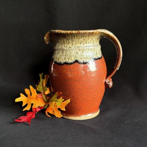 May include: A ceramic pitcher with a glossy, deep orange base and a textured, cream-colored rim featuring a dark drip pattern. The pitcher has a curved handle and a pouring spout, accompanied by a cluster of artificial orange, yellow, and green leaves.