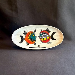 May include: An oval-shaped ceramic dish with a light gray wood-grain texture background. The dish features two stylized fish illustrations in vibrant orange, red, teal, and black, with yellow accents and a small black star-like shape between them.