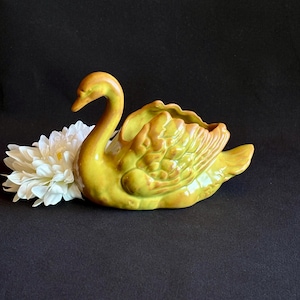 May include: A ceramic swan-shaped planter featuring a glossy chartreuse and golden-brown glaze with detailed molded feather textures on the wings. A large, white artificial chrysanthemum flower is positioned next to the swan figurine.