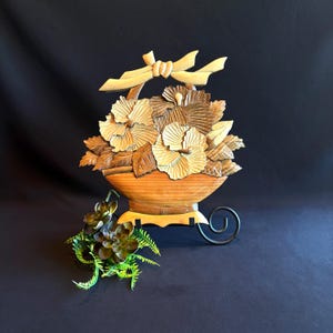 May include: A decorative wooden wall hanging featuring a carved basket filled with layered flowers and leaves, topped with a carved wooden bow. The piece is displayed on a black metal stand, accompanied by a small artificial green succulent plant.