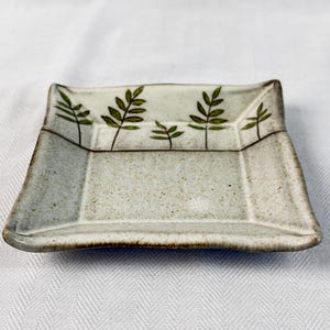 May include: A square ceramic dish with a speckled, off-white surface and a slightly raised rim. The dish is decorated with green leaf sprigs along the top edge. The dish is approximately 6 inches square.