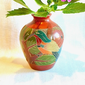 May include: A terracotta-colored ceramic vase featuring a hand-painted design of a bird with green, orange, and yellow plumage perched among green leaves and red fruit. The surface has a textured, matte finish with a smooth, rounded neck.