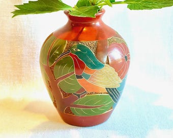 May include: A terracotta-colored ceramic vase featuring a hand-painted design of a bird with green, orange, and yellow plumage perched among green leaves and red fruit. The surface has a textured, matte finish with a smooth, rounded neck.