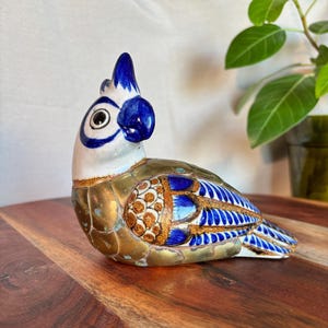 May include: A ceramic parrot figurine with a white head and blue crest, eyes, and beak. The body features gold and bronze tones with blue and orange patterned accents. The bird is perched on a wooden surface.