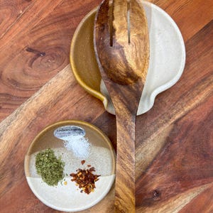 May include: A wooden salad fork rests on a two-toned ceramic spoon rest. A small ceramic dish holds herbs, salt, and red pepper flakes. The items are arranged on a wooden surface. The dish has the text "Lindsee Clements Handmade Ceramics".