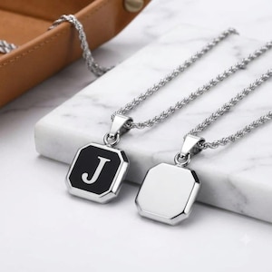 May include: Two silver-toned rope chain necklaces with octagonal pendants. One pendant features a black enamel inlay with the white letter J, while the other pendant is a plain, polished silver-toned metal surface without any markings or designs.