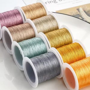 May include: A collection of spools of metallic thread in various colors, including pink, gold, silver, and orange. The thread is tightly wound on white plastic spools, creating a visually appealing display. The threads have a shiny, reflective quality.