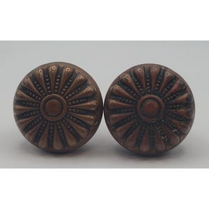 May include: Two antique bronze-colored door knobs with a floral design. Each knob features a central circle surrounded by a pattern of radiating petals and small dots. The knobs have a slightly aged appearance.