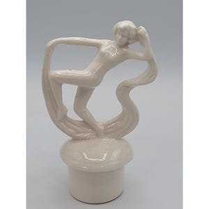 May include: A cream-colored ceramic bottle stopper featuring a stylized figure in a graceful pose. The figure appears to be dancing or reaching, with flowing lines suggesting movement. The stopper has a mushroom-shaped base.